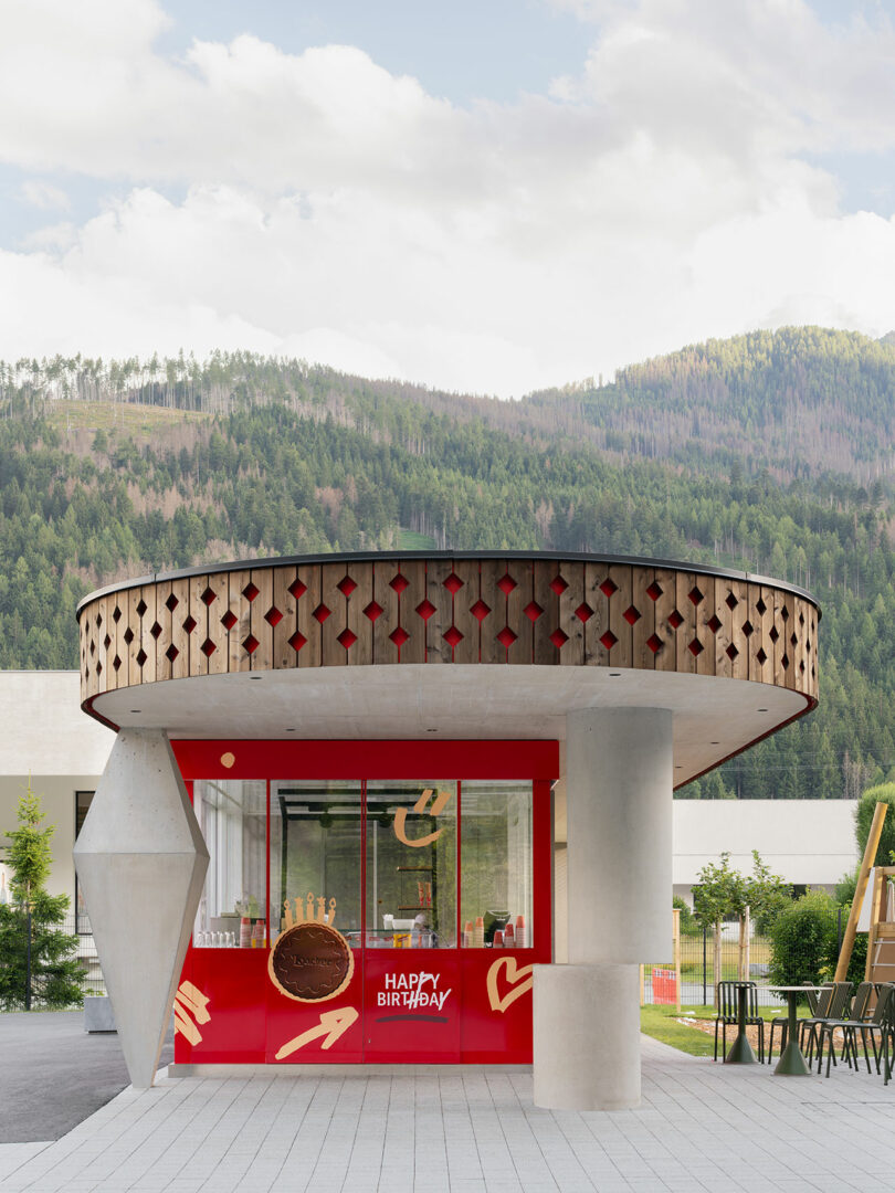 A small, modern red kiosk with geometric shapes and a decorative circular canopy is set against a backdrop of trees and mountains.