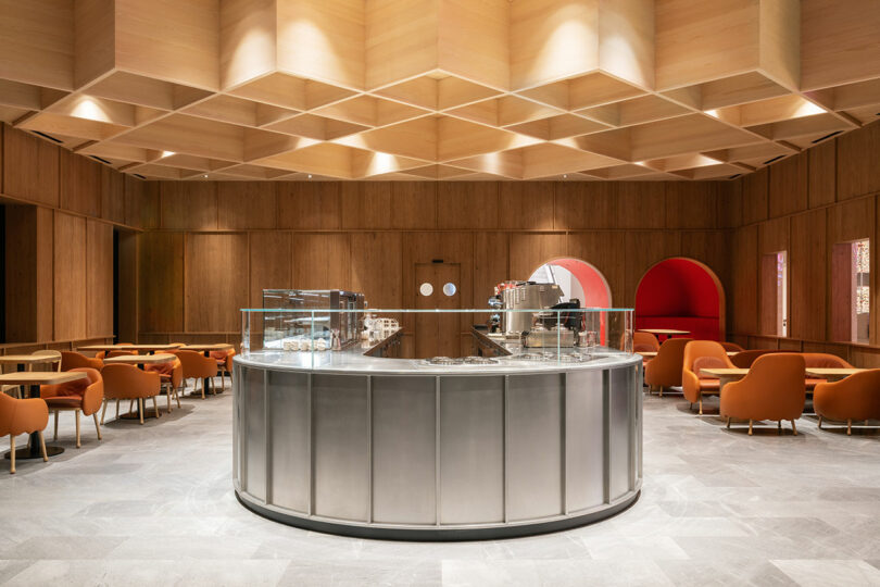 Modern café interior with a central circular metal counter, orange chairs, wooden paneled walls and ceiling, and geometric patterned design elements.