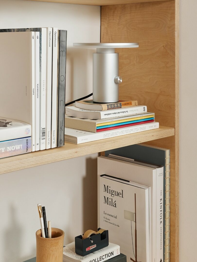 A modern bookshelf with neatly stacked books, a small silver lamp, a pencil holder with pens, and a tape dispenser on wooden shelves