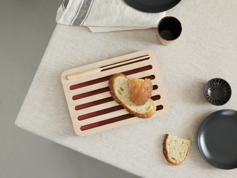 A wooden slotted breadboard with a slice of bread on it sits on a beige tablecloth next to a black plate, a cup, and a small bowl. Another bread slice is on the table