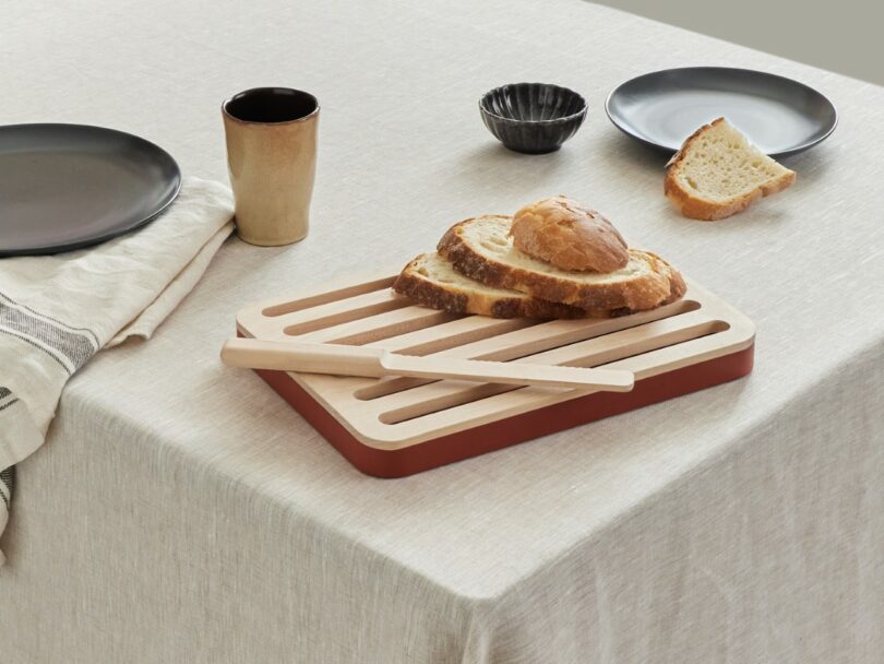 Sliced bread on a wooden cutting board with tongs, next to plates, a cup, and a striped cloth on a beige tablecloth