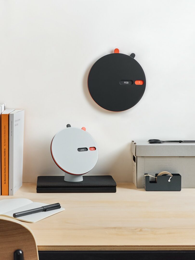 Two round, modern wall clocks—one black, one white—are displayed on a desk and wall above an organized workspace with books, boxes, and office supplies