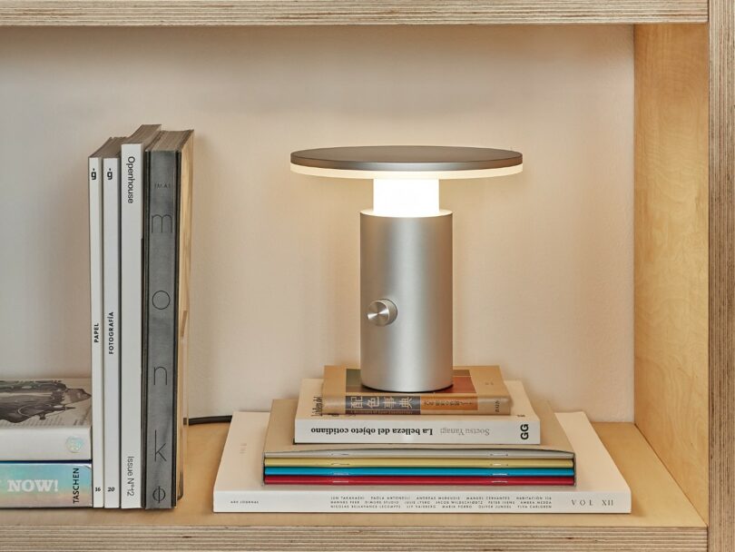A modern silver table lamp sits on a stack of books inside a wooden bookshelf, surrounded by vertically and horizontally arranged books