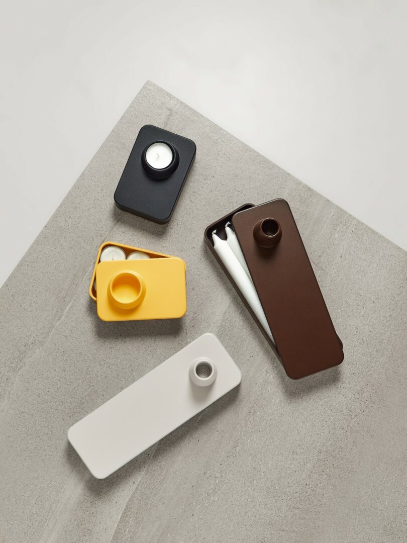 Rectangular plastic containers in black, yellow, brown, and white with rounded corners and circular lids, arranged on a light gray textured surface