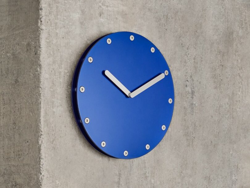 A blue wall clock with silver hour markers and hands, displaying the time as 10:10, mounted on a concrete wall