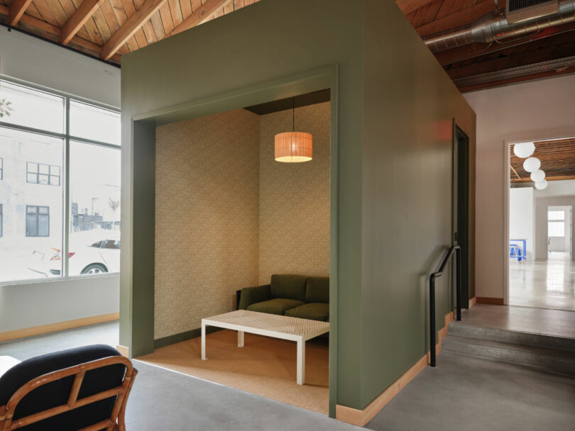 A small seating nook with a green sofa, white coffee table, and a hanging light is built into a green-walled cubicle inside a modern office space with large windows and exposed ceiling.
