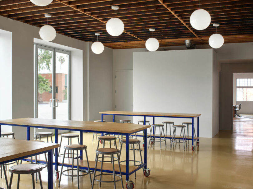 A spacious room with high tables and stools on casters, light fixtures on a wooden ceiling, and large windows letting in natural light.