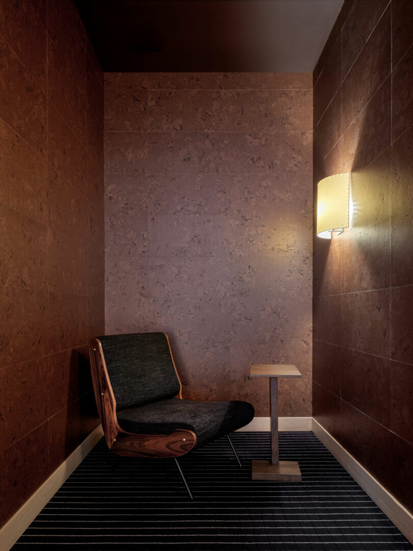 A single cushioned chair and small wooden table sit against brown textured walls under a wall-mounted lamp in a minimalist, modern corner space with striped carpet flooring.