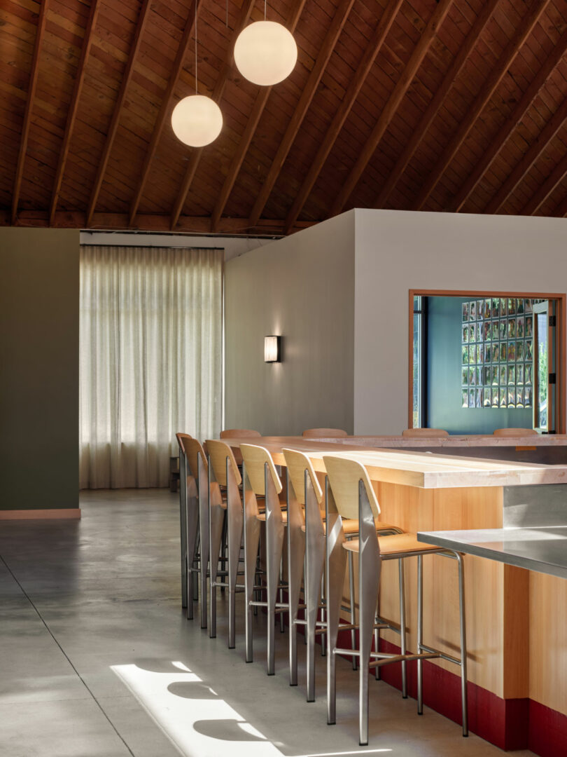 A modern interior with a wooden ceiling, pendant lights, a long bar counter with wooden stools, concrete floor, and a curtained window.