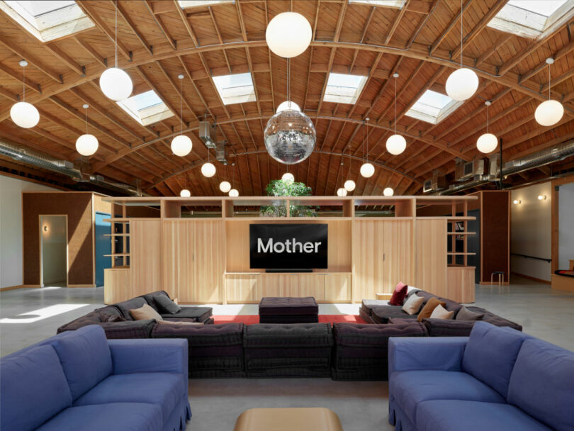 Spacious lounge with blue and black sofas, wooden ceiling, globe lights, a disco ball, and a large screen displaying "Mother.