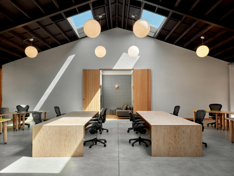 A modern open office with long wooden desks, black office chairs, skylights, large round ceiling lights, and a private seating area at the back.