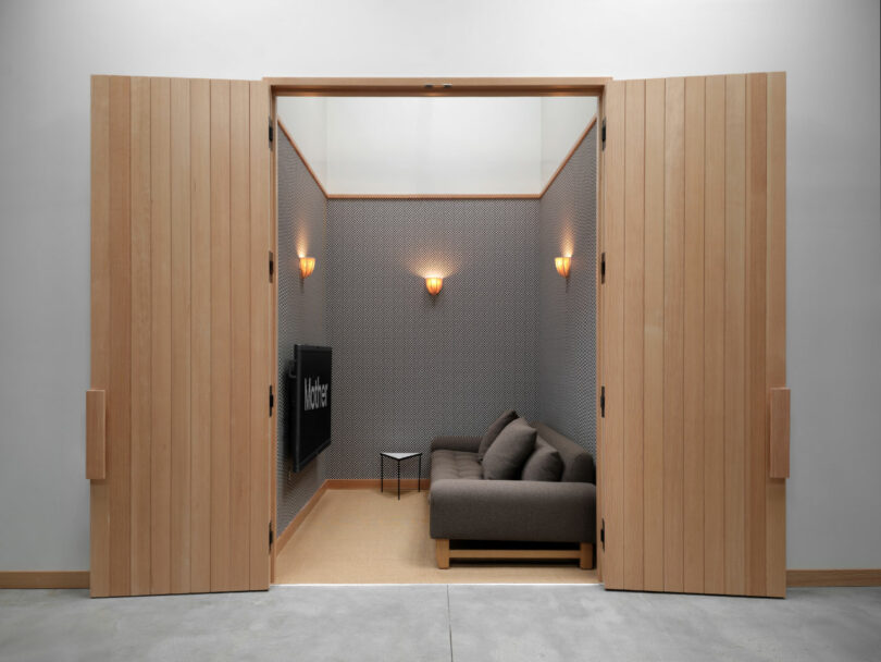 A small room with wooden double doors, a gray sofa, a wall-mounted TV, a small side table, and two wall sconces.