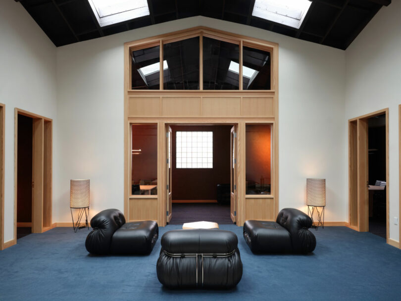 Modern lounge area with three black leather chairs, a low white table, blue carpet, tall lamps, and wooden-framed glass partitions beneath a vaulted ceiling with skylights.