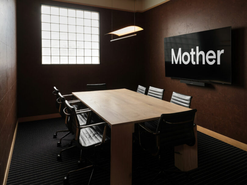 A conference room with a rectangular table, seven black chairs, a large window, and a wall-mounted screen displaying the word "Mother.