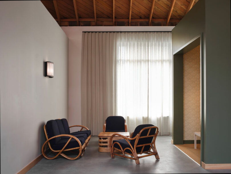 A minimalist room with a wooden ceiling, neutral walls, sheer curtains, and rattan furniture including two chairs, a sofa, and a coffee table.
