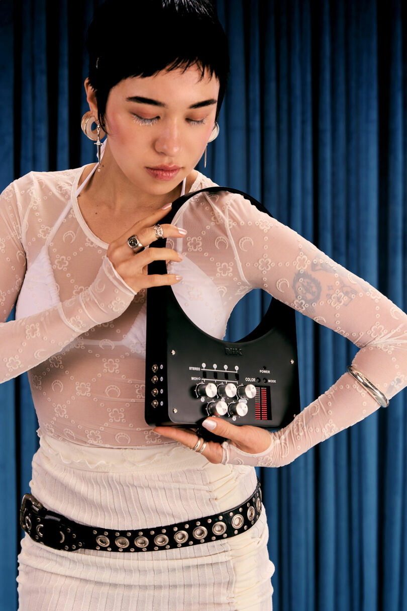 A person with short hair stands in front of blue curtains, holding a black electronic device with knobs and switches, wearing a sheer white top and studded accessories.