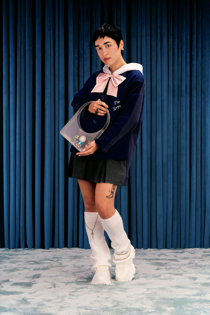 A person with short dark hair stands in front of blue curtains, wearing a navy sweater with a pink bow, a skirt, knee-high white socks, and white shoes, holding a modern circular device.