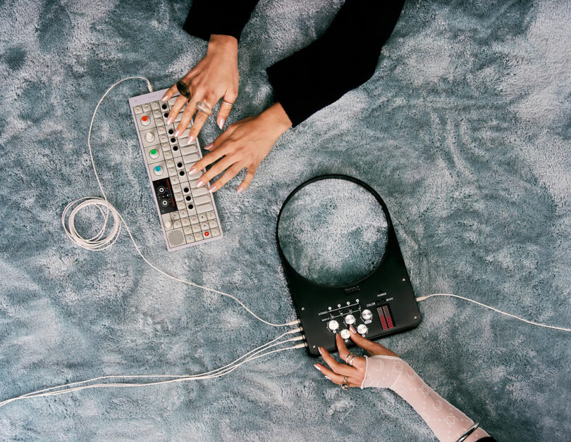 Two people’s hands operate electronic music devices on a textured blue surface, with cords connecting the equipment.