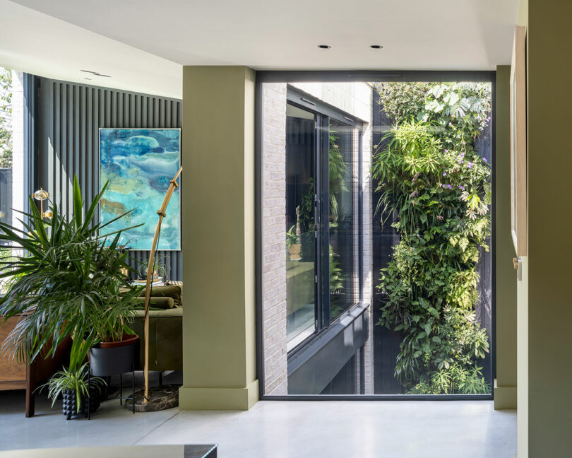 Modern interior with green walls, large window, indoor plants, a floor lamp, and a view of an outdoor vertical garden.