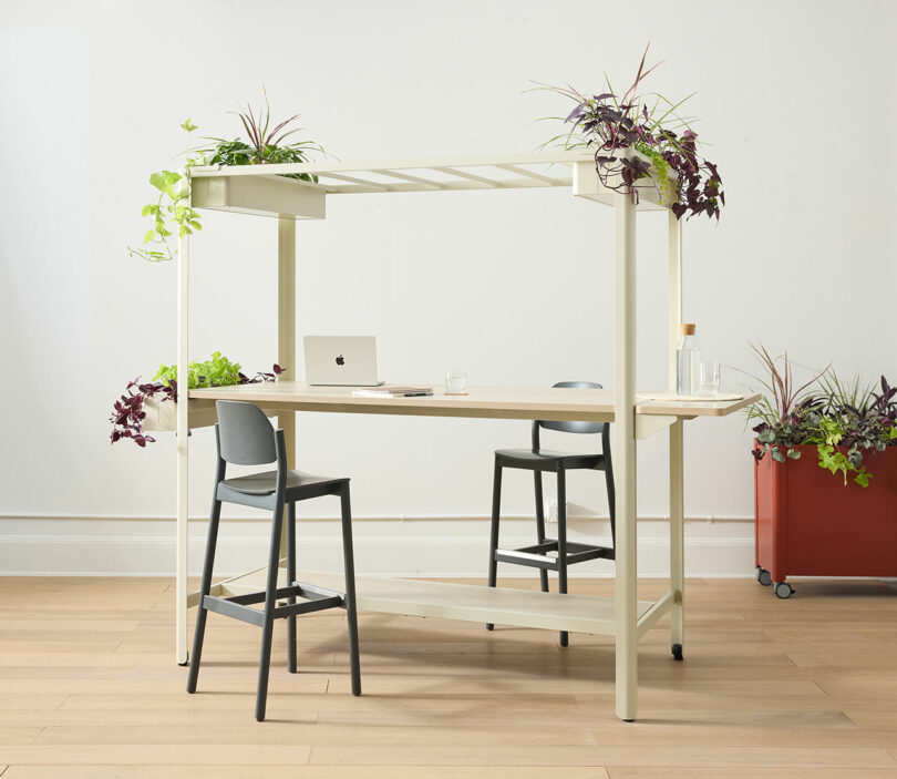A modern desk with plants on top and around the sides, two chairs, a closed laptop, a notebook, a bottle, and a glass in a bright room with wooden flooring.