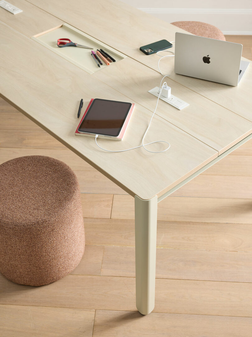 A light wood desk with two stools, a laptop, tablet, phone, pens, scissors, and charging cables organized on the surface.