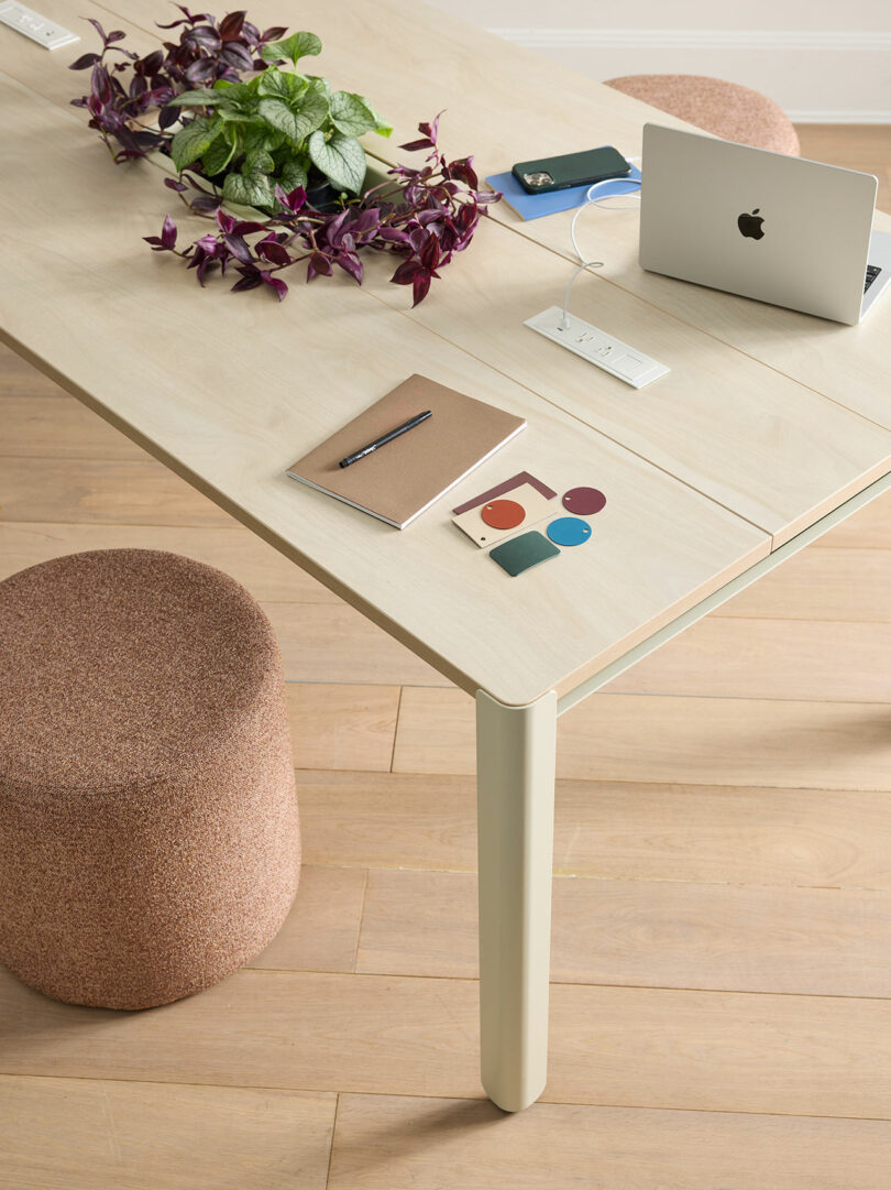A light wood table with a closed laptop, notebook, pen, a plant, color swatches, and a charging station; a brown stool is nearby on a wooden floor.