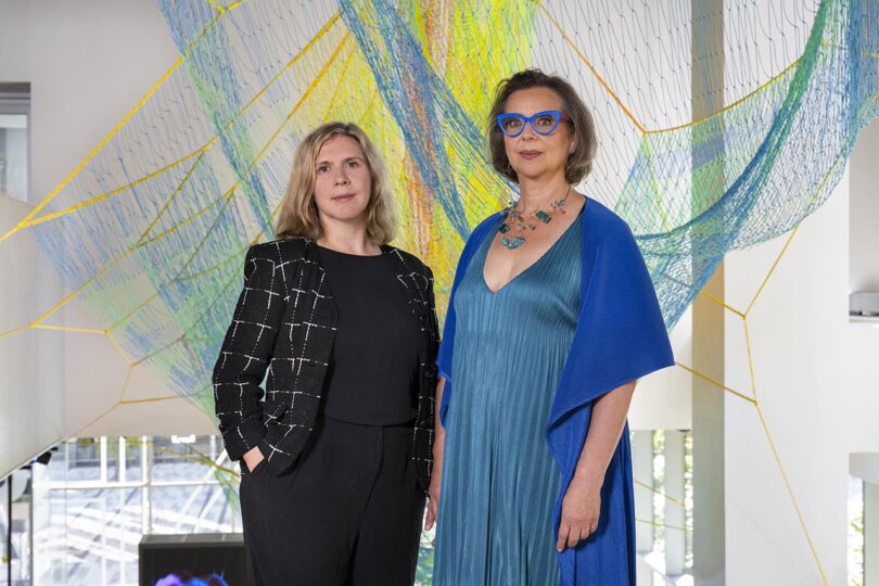 Two women stand indoors in front of colorful net art installation; one wears a black jacket and pants, the other a blue dress and shawl with blue glasses.