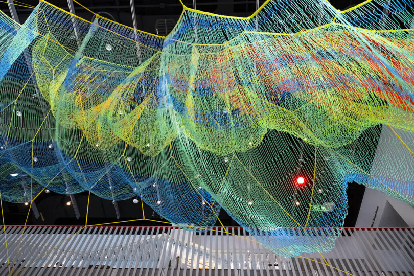 A colorful net sculpture made of string is suspended from a ceiling, with lights and architectural details visible in the background.