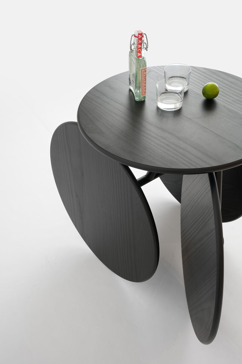 A round black table with uniquely shaped legs, featuring a glass bottle, two empty glasses, and a lime on its surface.