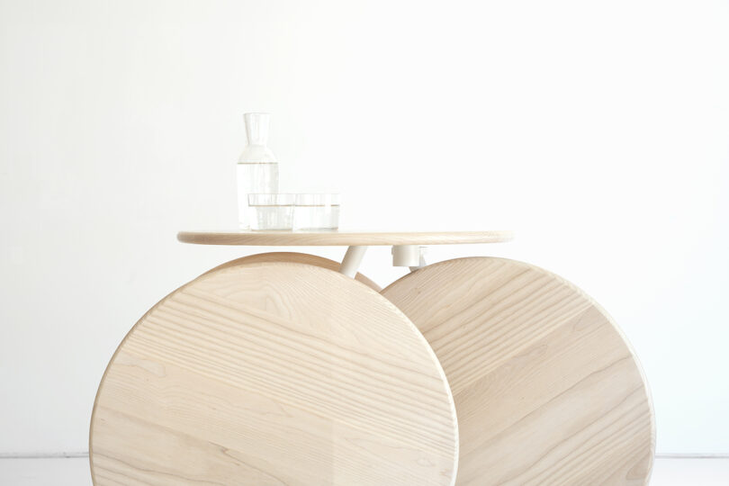 A minimalist wooden table with two large circular legs holds a glass carafe and two drinking glasses, set against a plain white background.
