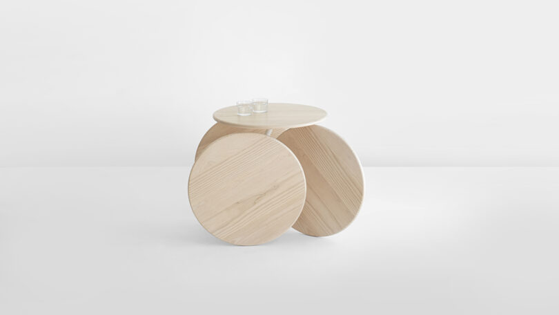A modern wooden table with two large circular legs and a round tabletop, holding two glass cups, set against a plain white background.