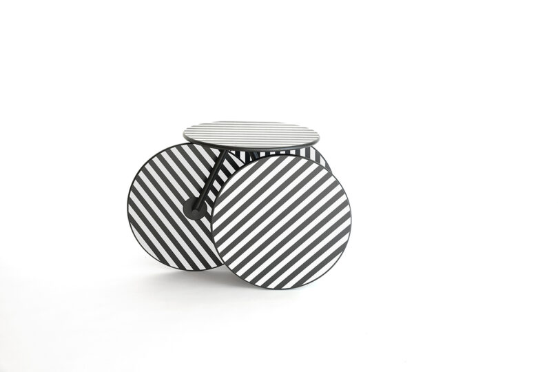 A small table with three large, flat circular legs and a round tabletop, all featuring black and white diagonal stripes, set against a plain white background.