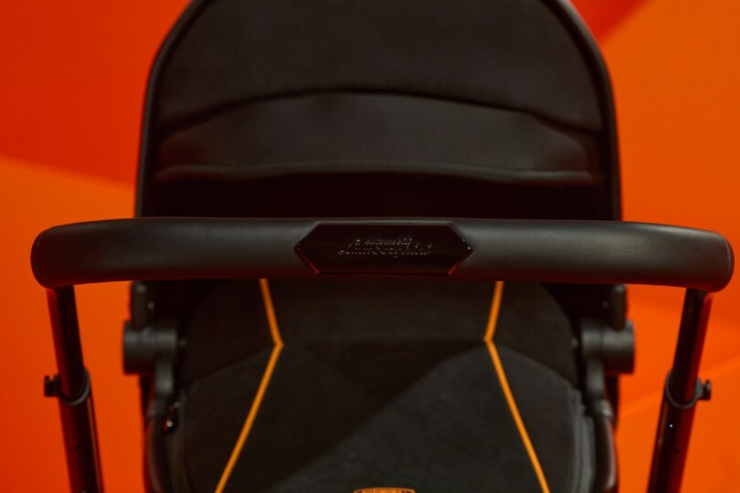 Close-up of a Silver Cross Lamborghini black stroller handle with stitching detail against an orange background.