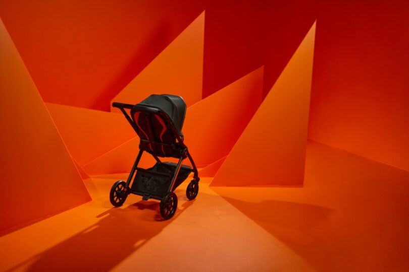 A Silver Cross Lamborghini black stroller is positioned in a bright orange room with geometric, angular wall structures