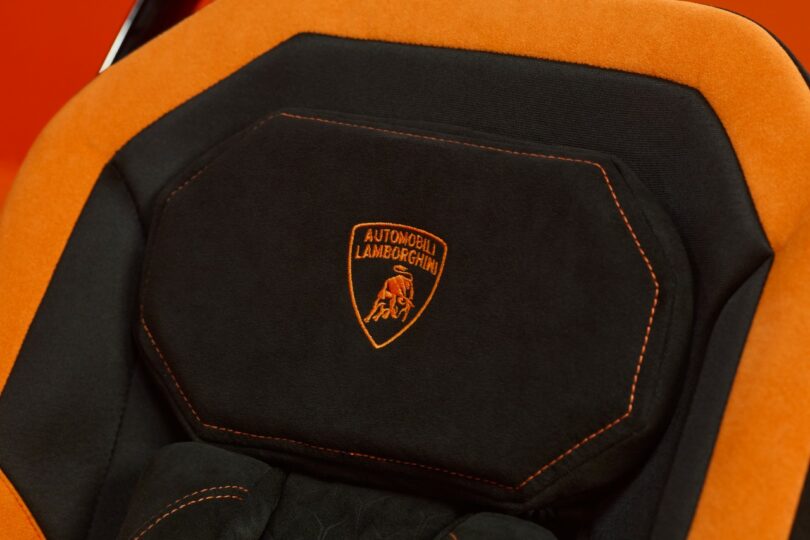 Close-up of a car seat with black and orange upholstery, featuring the Lamborghini logo and "Automobili Lamborghini" embroidered in orange thread—a signature detail of the Silver Cross Lamborghini collection