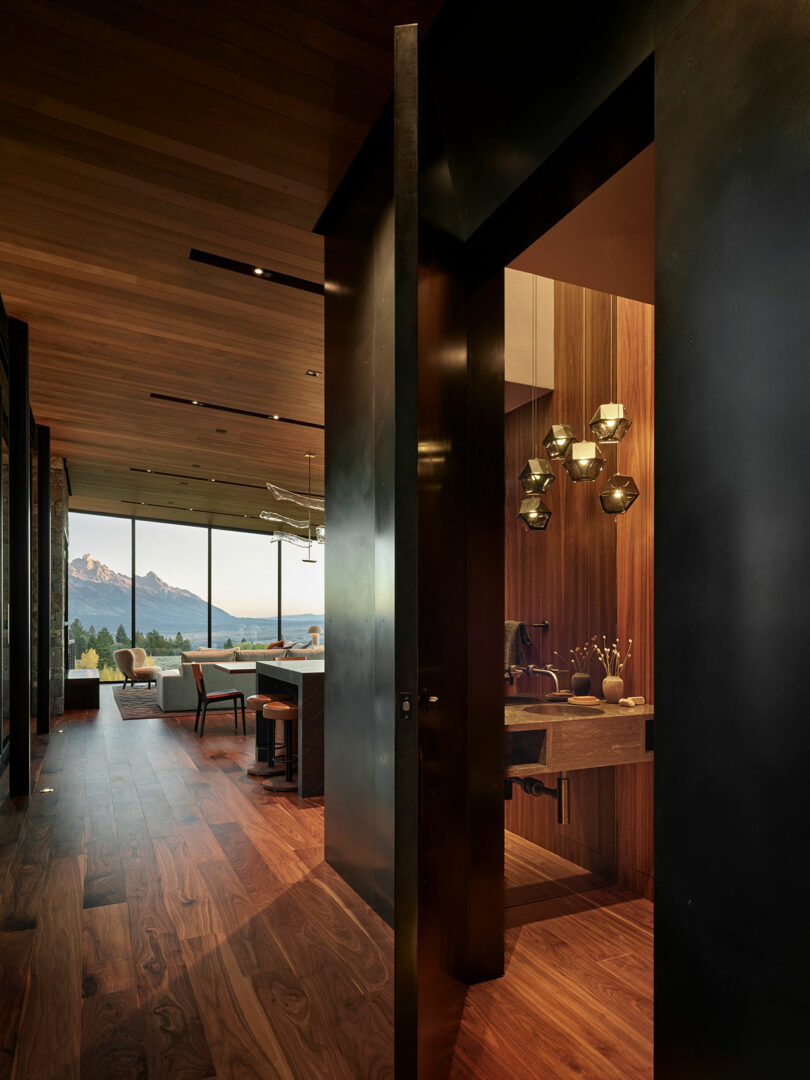 Modern interior hallway with wood flooring and ceiling, open to a living area with mountain views; a bathroom with pendant lights is visible to the right.