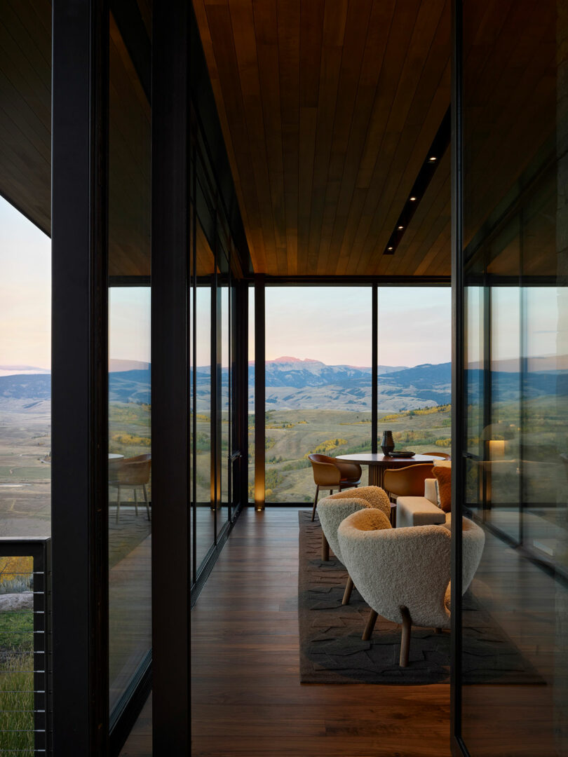 A modern glass-walled room with two armchairs and a small table overlooks a scenic mountain landscape at sunset.