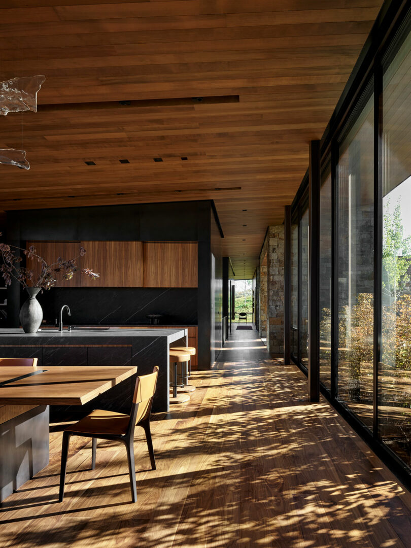 Modern kitchen and dining area with wood floors, wood ceilings, dark cabinets, large windows, and sunlight casting shadows from trees outside onto the interior space.