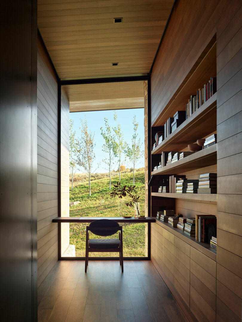 A narrow modern study with wood-paneled walls, built-in bookshelves, a desk, and a chair, featuring a large window overlooking a grassy landscape with trees.