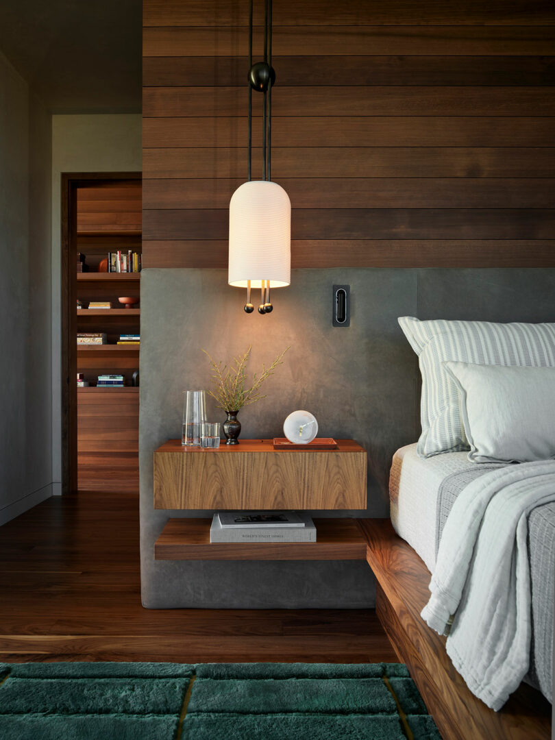A modern bedroom with a wooden accent wall, pendant light, floating nightstand holding a vase, glass, and clock, and a bed with striped bedding. A bookshelf is visible in the background.