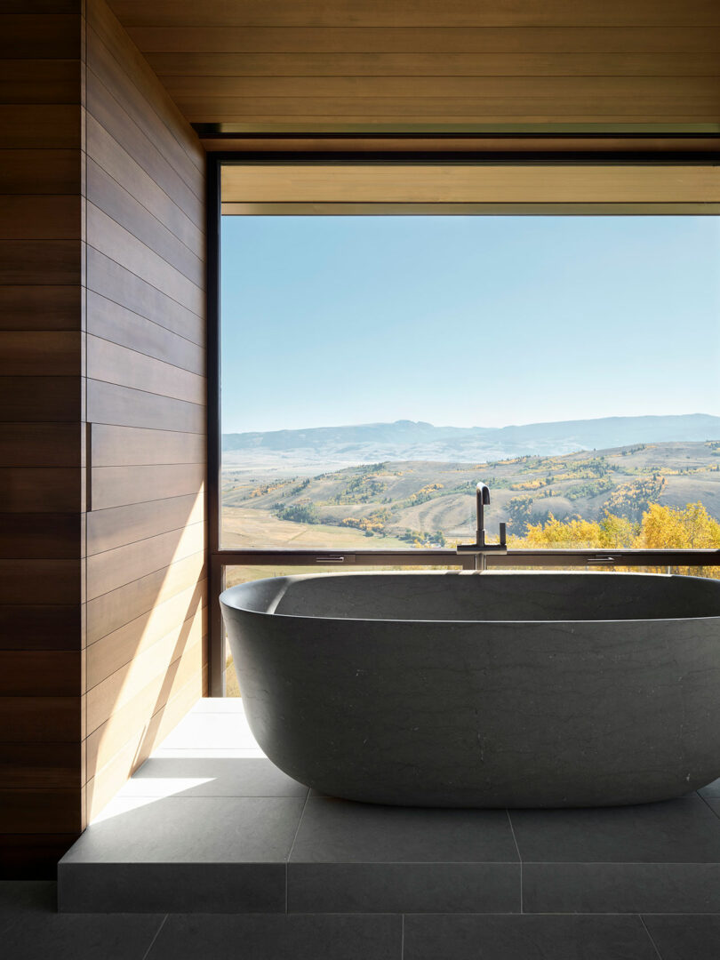 A modern freestanding black bathtub is positioned by a large window with a scenic mountain and valley view; the room features wood and tile surfaces.