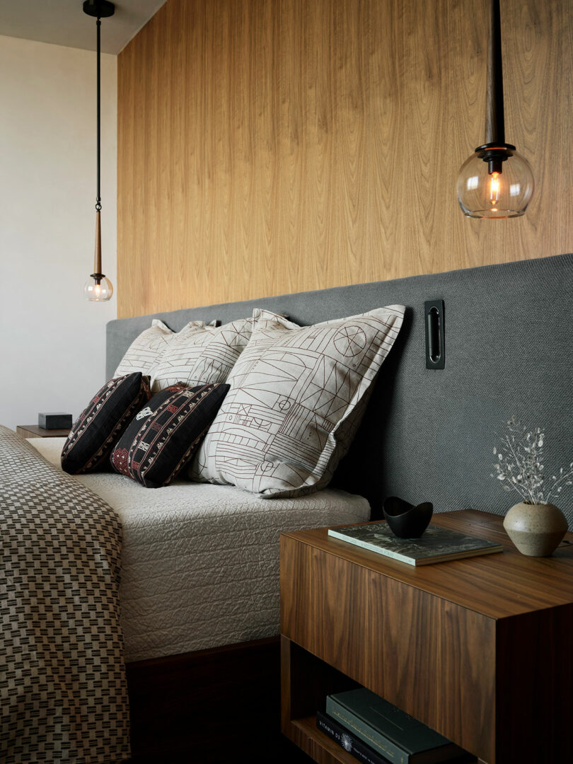 A modern bedroom with a gray upholstered headboard, patterned pillows, a wood nightstand with books and decor, and two hanging glass pendant lights.