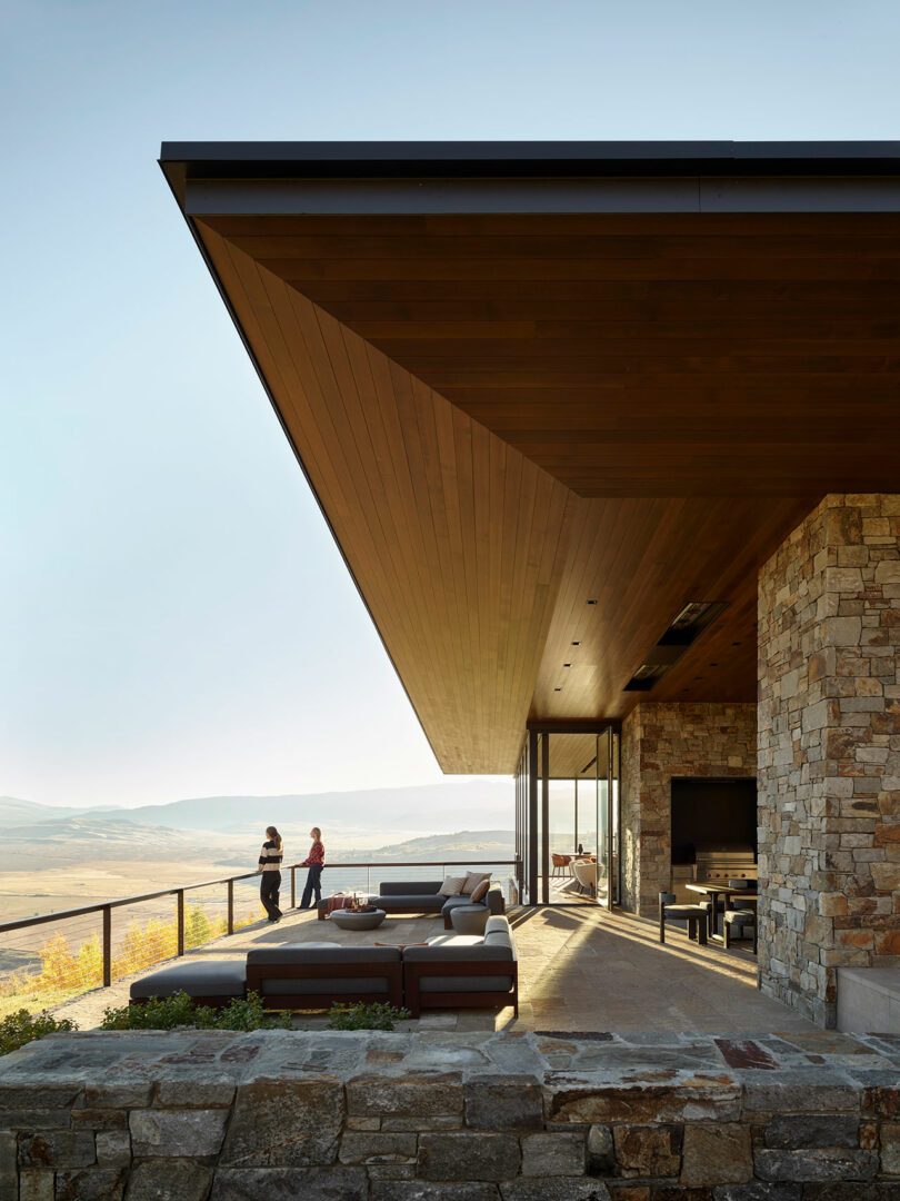 Modern house with stone walls and a large wooden overhang, outdoor seating area, and two people standing on a terrace overlooking a scenic landscape.