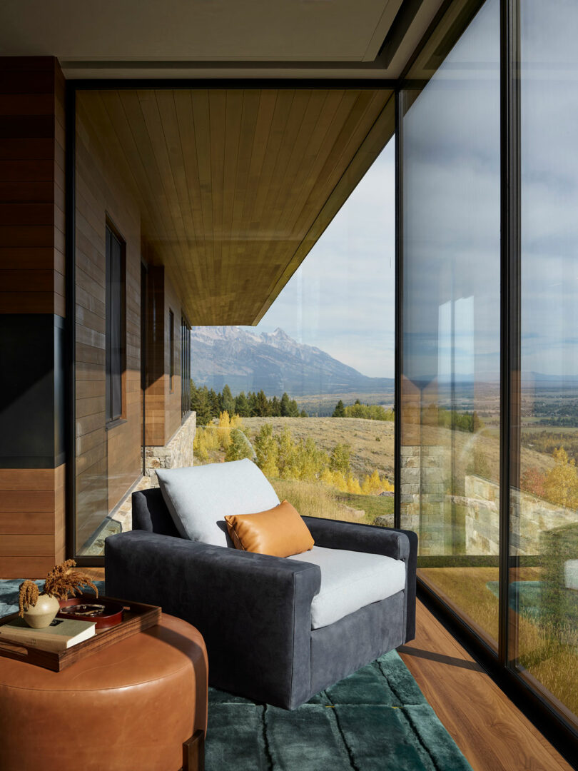 A modern lounge chair with a tan pillow sits by floor-to-ceiling windows, overlooking a scenic mountain and forest landscape.