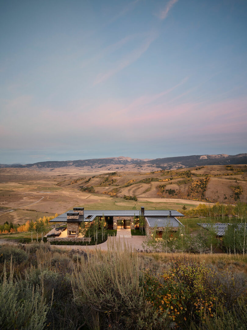 Modern house with large windows sits on a hillside surrounded by sparse vegetation, overlooking a wide valley and distant mountains under a pastel sky at dusk.