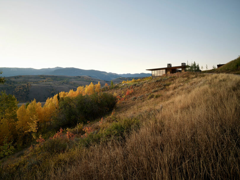 A modern house sits on a grassy hillside overlooking a valley with autumn trees and distant mountains under a clear sky.