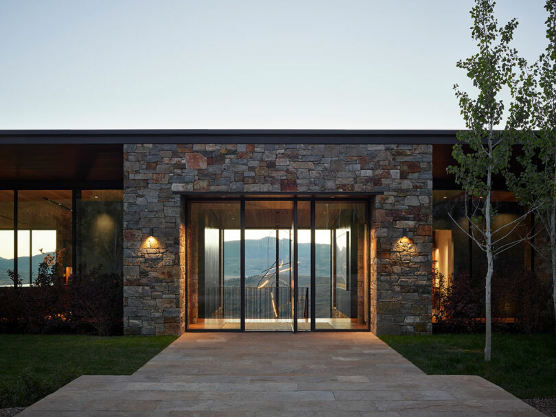 Modern house with a stone facade and large glass entrance, illuminated by exterior lights, with a tree and mountain view in the background.