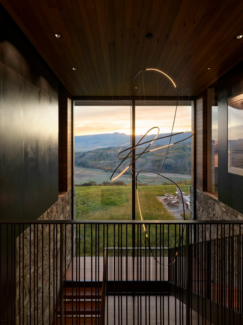 Modern interior with a wooden staircase, stone walls, and a sculptural light fixture, overlooking an outdoor patio and scenic landscape through large floor-to-ceiling windows at sunset.