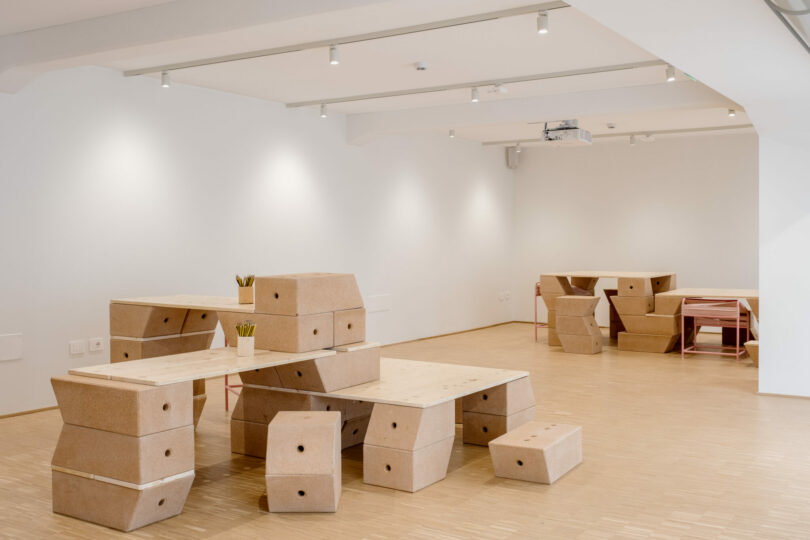 Minimalist room with light wooden floors, white walls, and modular cork and wood furniture arranged as desks, benches, and stools; some yellow pencils in holders on tables