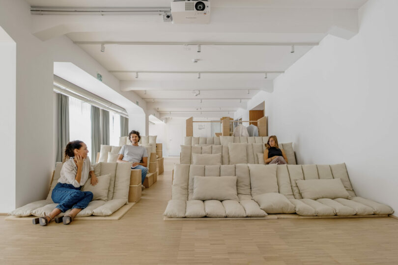 Three people sit on cushioned seating arranged in tiered rows in a bright, modern room with white walls and wooden floor
